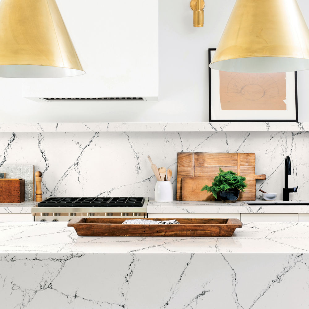 a modern kitchen with white veined quartz countertops and matching quartz island and backsplash, with two pendant light fictured, a sleek hood, and white cabinets. 
