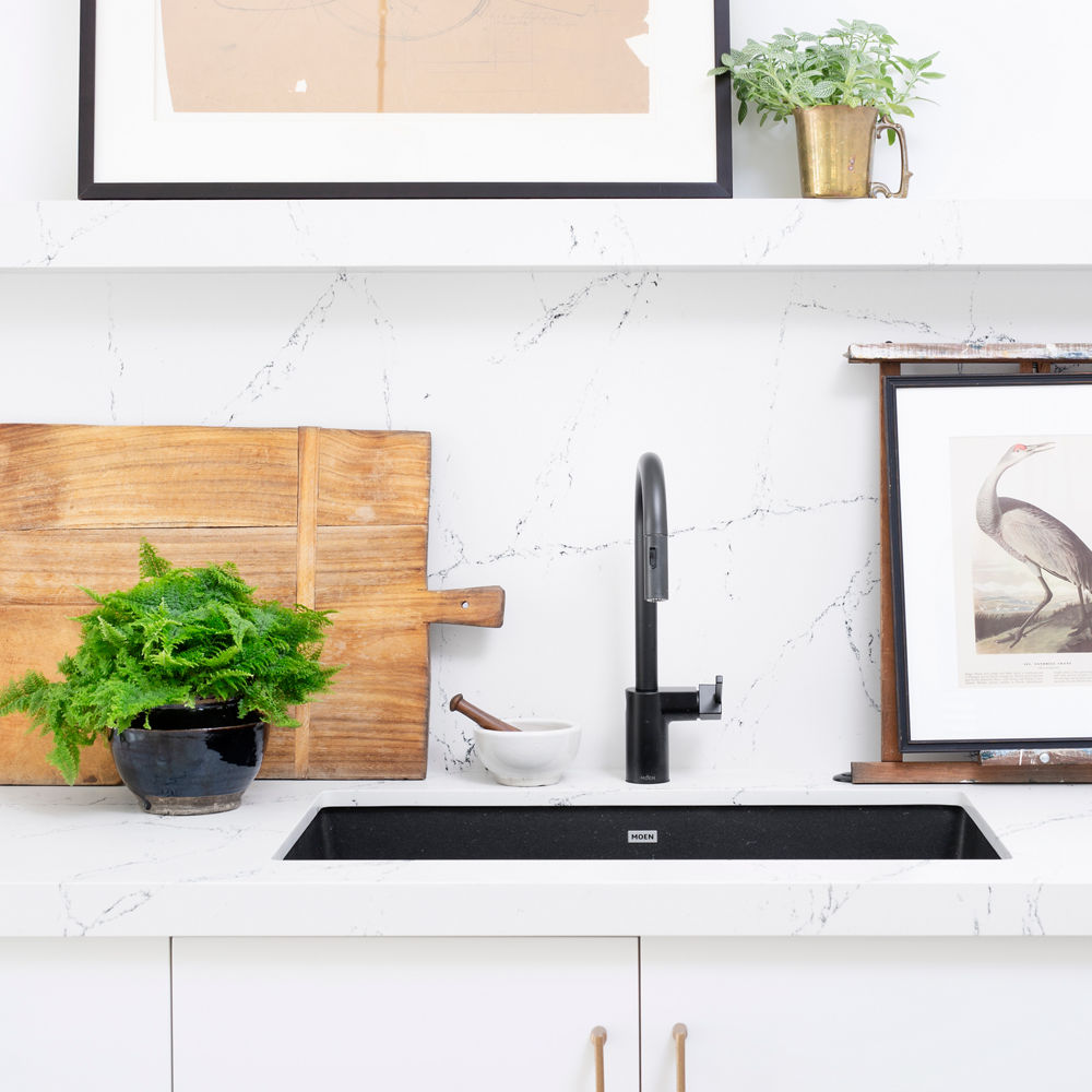 a modern kitchen with a black sink, white cabinets, white quartz countertops and matching backsplash, and gold accents.