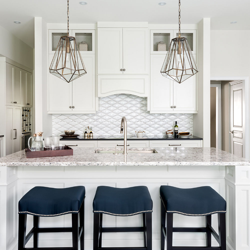 A white kitchen with Cambria Bellingham quartz kitchen island featuring three navy blue bar stools and eye-catching gold light fixtures.