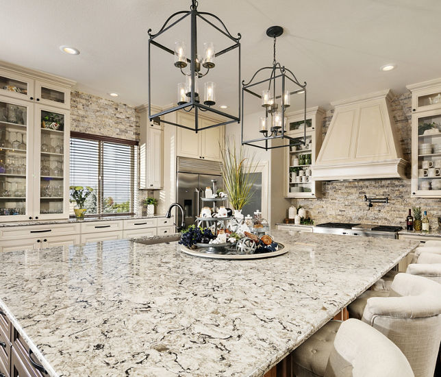 a traditional kitchen with tan upper and lower cabinets, large white hood over range large center island topped with quartz countertops, with pendant lights and barstools