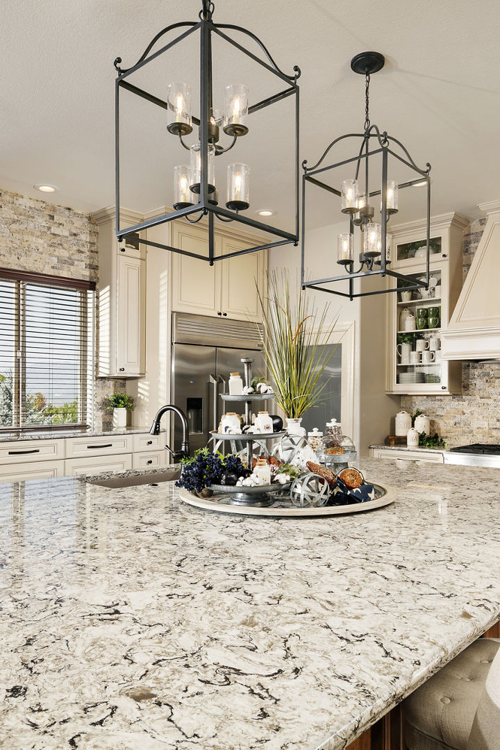a traditional kitchen with tan upper and lower cabinets, large white hood over range large center island topped with quartz countertops, with pendant lights and barstools