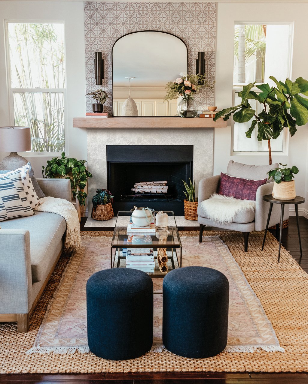 a warm and inviting living room with a quartz fireplace, mirror above the mantle, fun wallpaper, couches with multiple pillows and blankets, and a fun rug. 