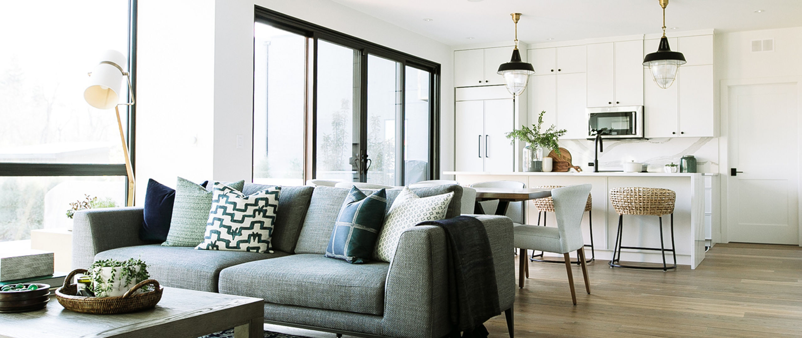 A cozy living room with a gray couch in front of a coffee table in front of it. The open kitchen, dining room, and living room are connected beside a bright window.