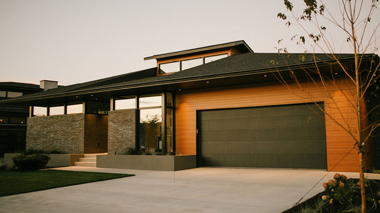 A house with black garage door surrounded by wooding with a white driveway and entryway, with windows giving a glimpse into the interior.