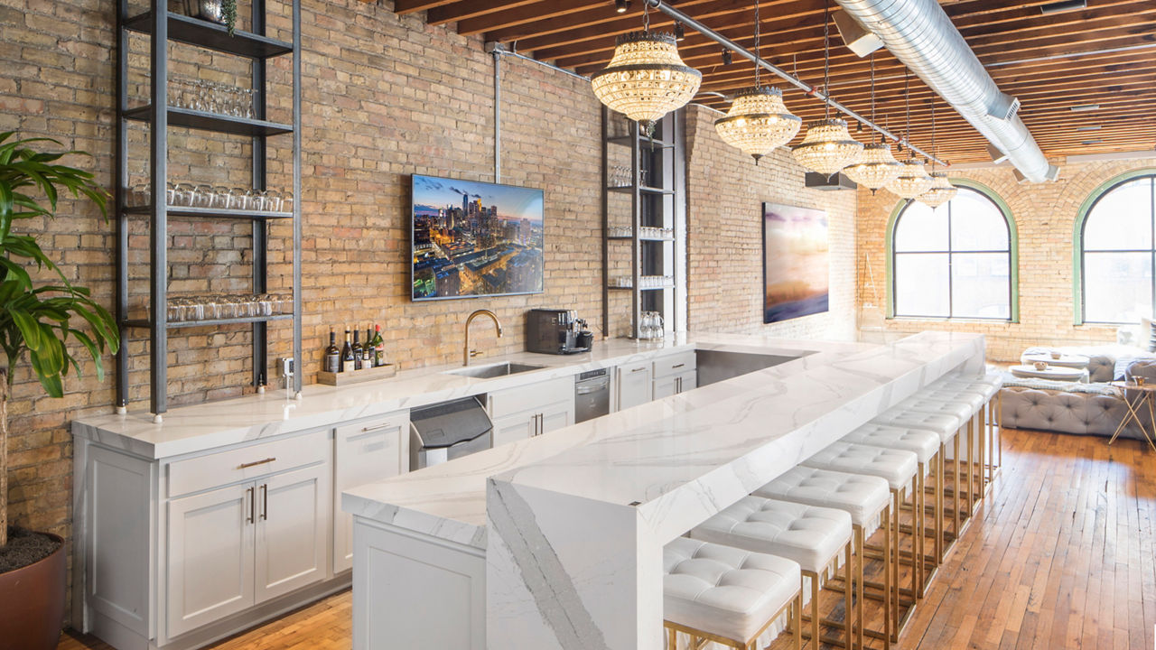 an industrial style bar with a bar with white cabinets, white quartz countertops and bar seating area, white and gold bar stools, and open shelving. 