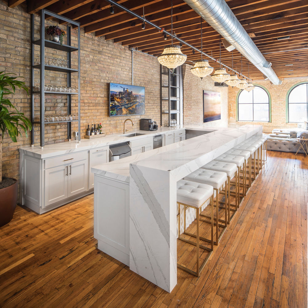 an industrial style bar with a bar with white cabinets, white quartz countertops and bar seating area, white and gold bar stools, and open shelving. 