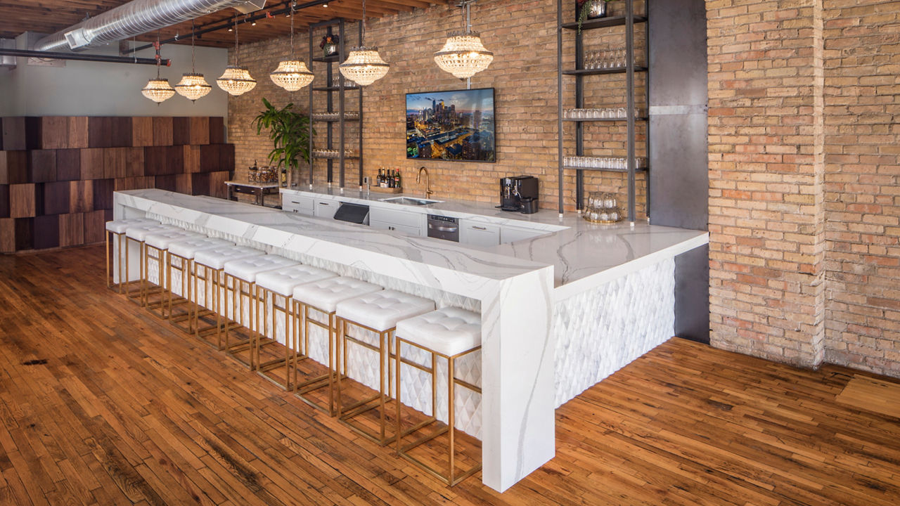 A white bar counter with ten stools sitting in front of the counter surrounded by wooden flooring and brick siding.