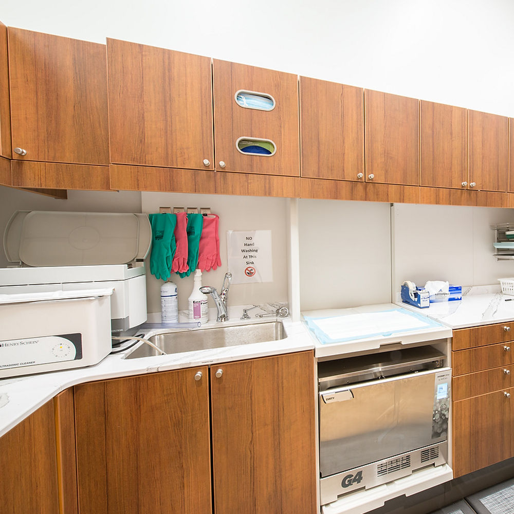 A dental office cleaning room with upper and lower oak cabinets, white quartz countertops, a gray rug, and various cleaning equipment