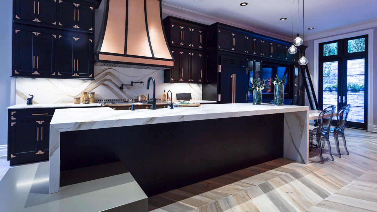 A kitchen with dark blue lighting and dark cabinets with a Brittanicca Gold Warm waterfall countertop