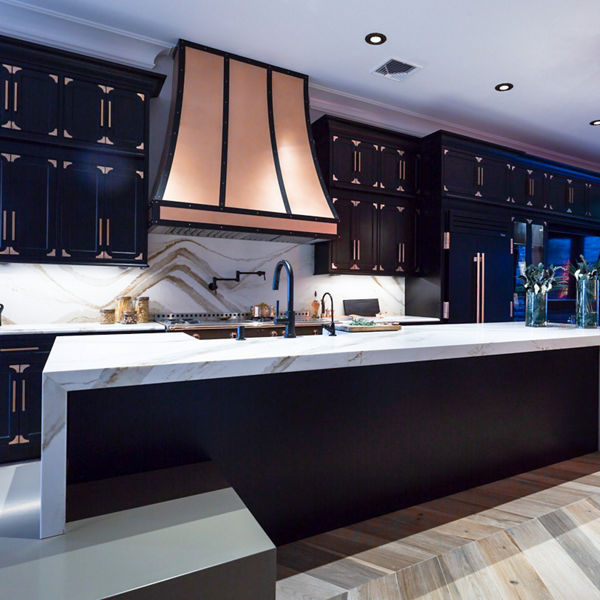 A kitchen with dark blue lighting and dark cabinets with a Brittanicca Gold Warm waterfall countertop