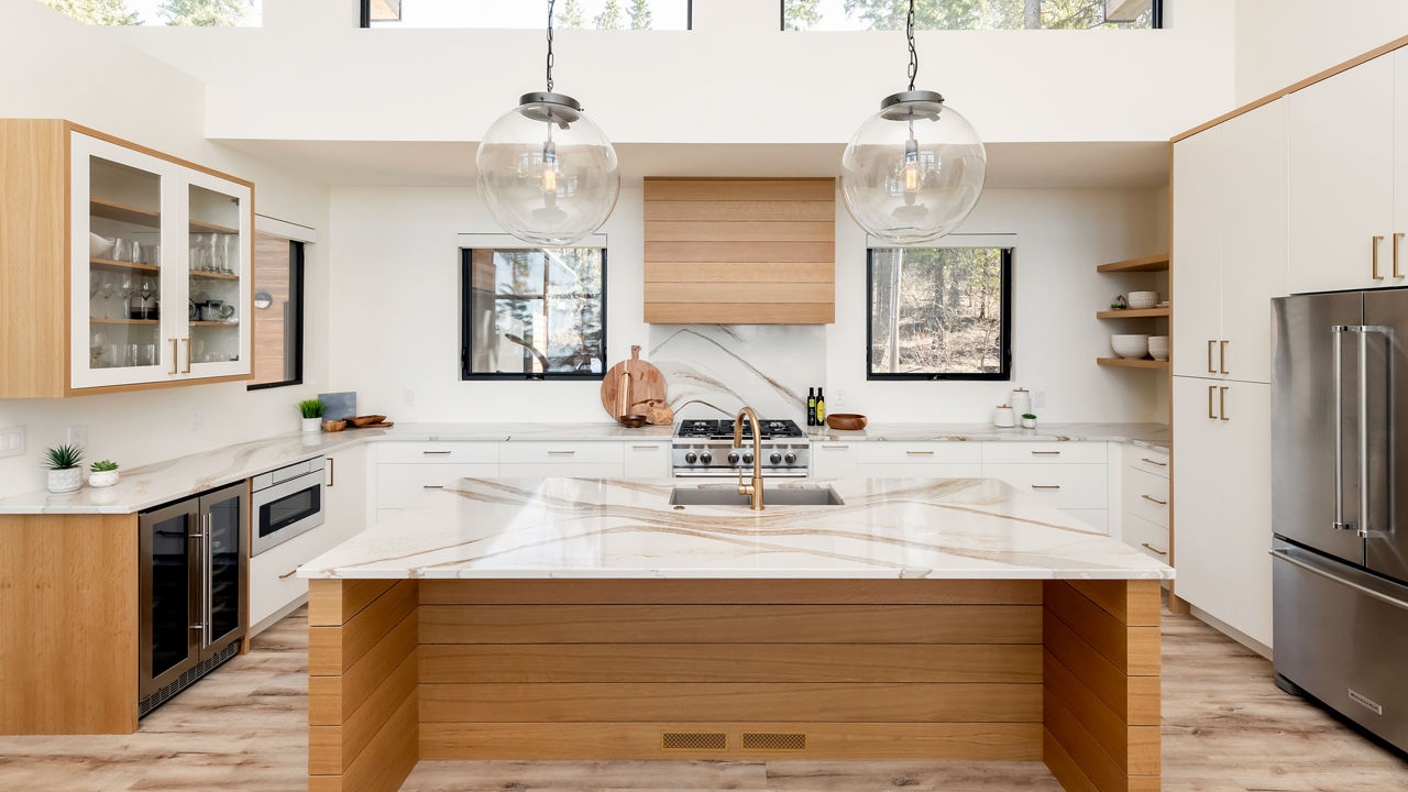 a modern kitchen with plenty of natural lighting, wooden cabinets and island topped with quartz countertops, two bulb lights hanging over the island, and a wooden hood over the range.