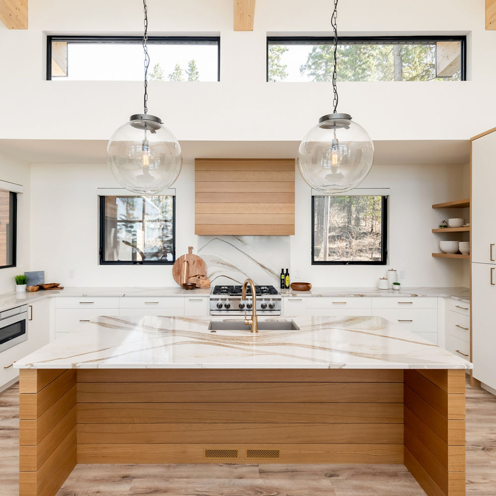 a modern kitchen with plenty of natural lighting, wooden cabinets and island topped with quartz countertops, two bulb lights hanging over the island, and a wooden hood over the range.