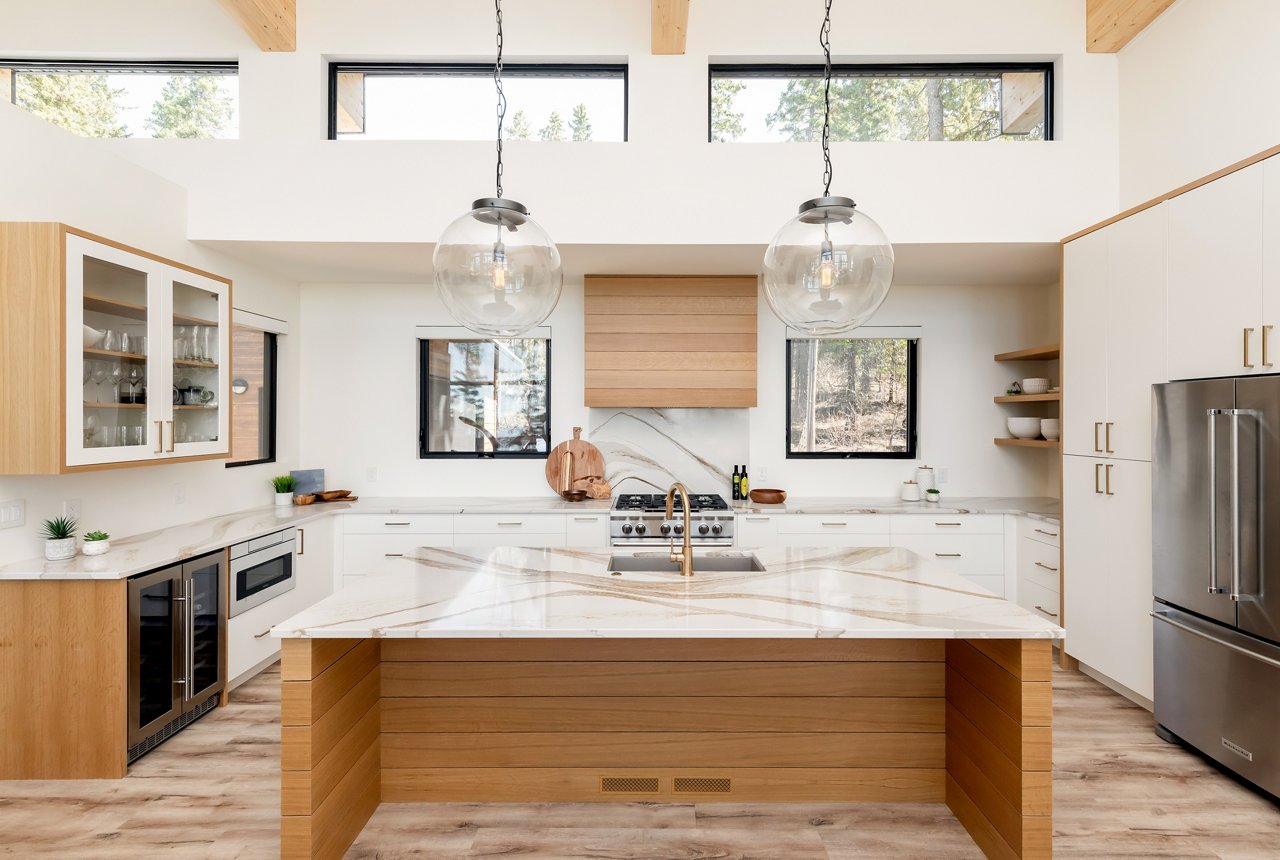 a modern kitchen with plenty of natural lighting, wooden cabinets and island topped with quartz countertops, two bulb lights hanging over the island, and a wooden hood over the range.