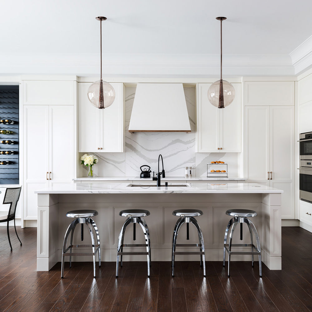 A white kitchen with white cabinets, white quartz countertops and matching quartz backsplash, white hood over stove, center island with quartz countertops and built in sink and 4 barstools with a dining table to the left with a built-in wall wine rack