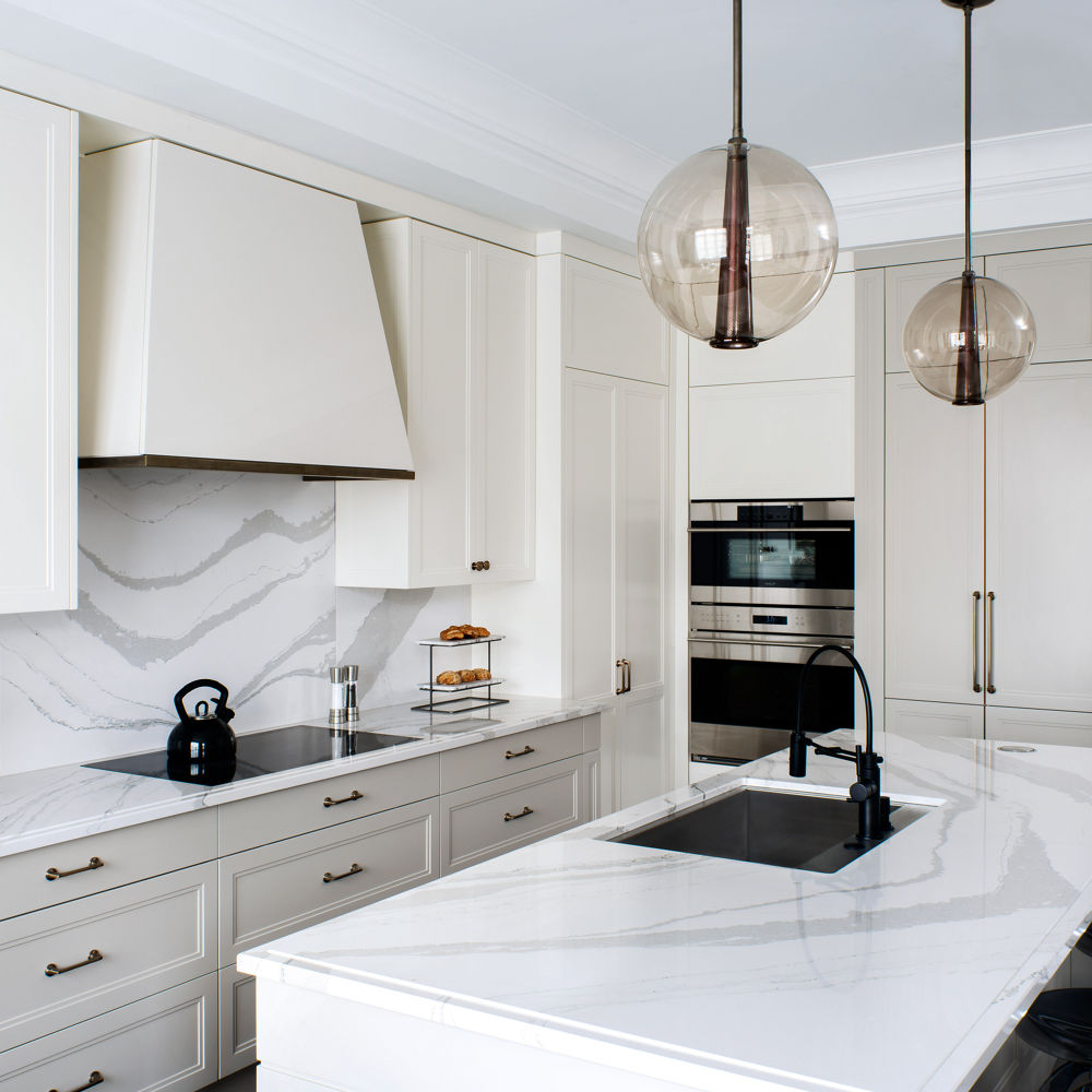 A white kitchen with white cabinets, white quartz countertops and matching quartz backsplash, white hood over stove, center island with quartz countertops and built in sink and 3 barstools