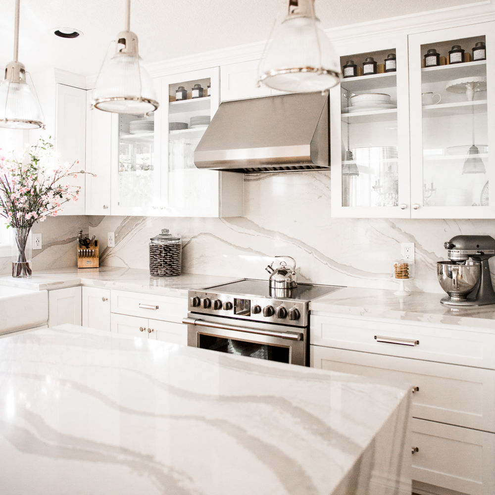 A clean white kitchen with white cabinets, white quartz countertops, stainless steel appliances, lots of natural lighting and overhead pendant lights.