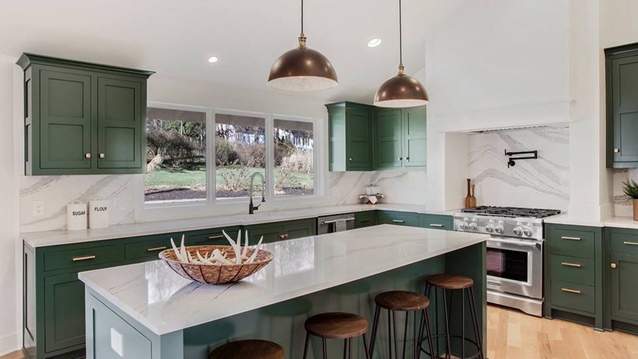 A kitchen with emerald green cabinets, white quartz countertops, bronze accents, and wood and metal barstools.
