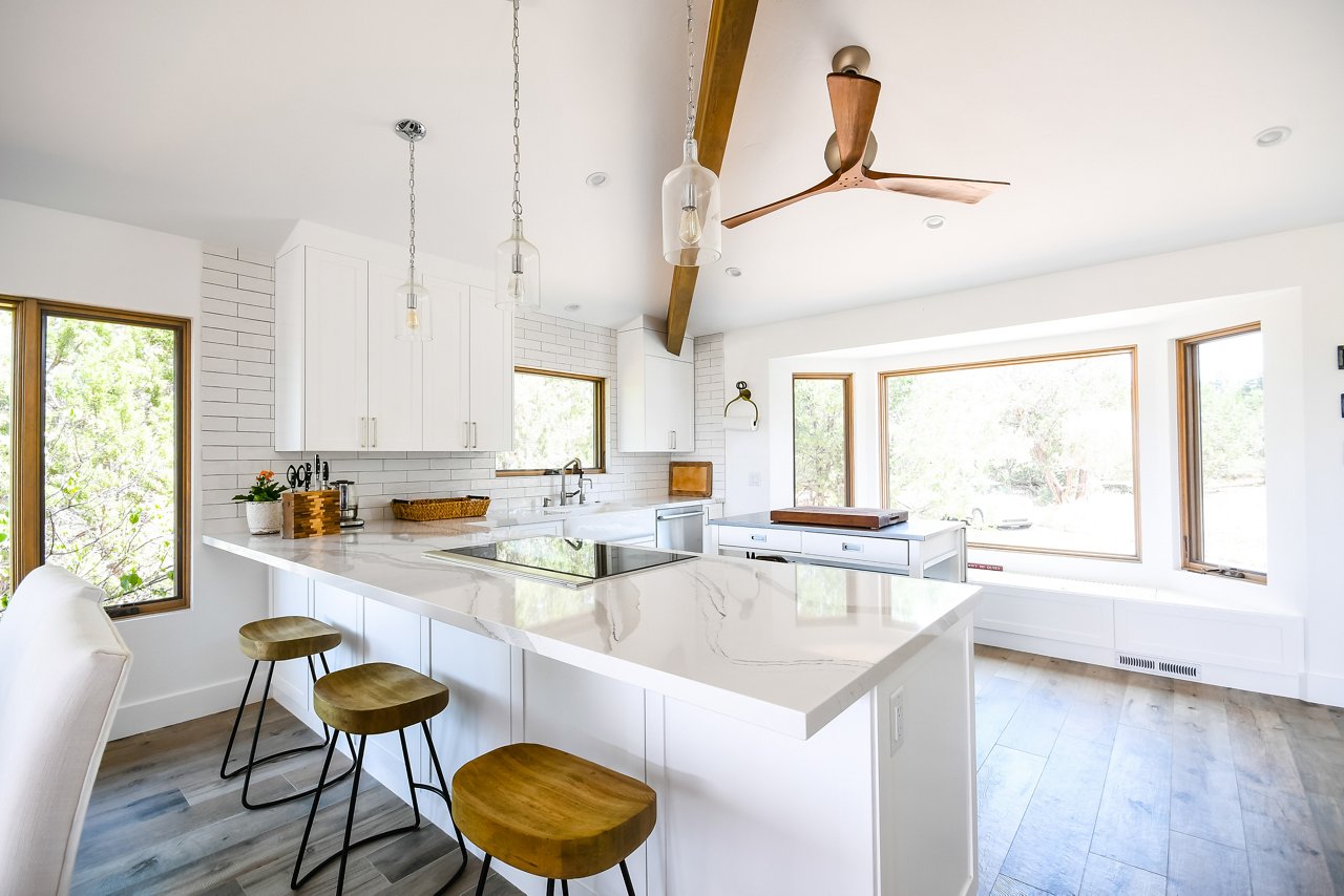 A simple white kitchen with high ceilings, lots of natural light, pendant lighting over the island, wooden accents and barstools, and Brittanicca quartz countertops.