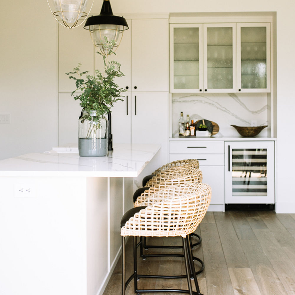 a clean kitchen with a white island topped with white quartz countertops, whicker style bar stools, and a dry bar with small wine refrigerator and quartz backsplash.