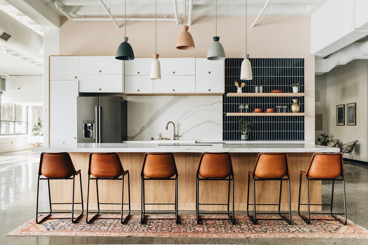 A modern kitchen with white quartz countertops and matching quartz backsplash, white cabinets, open shelving with black tile backsplash behind it, and six leather barstools