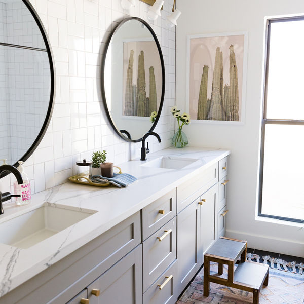 A bathroom with a gray vanity with two sinks, white quartz countertops, two black circular mirrors, gold wall lights above the mirrors, and a step stool