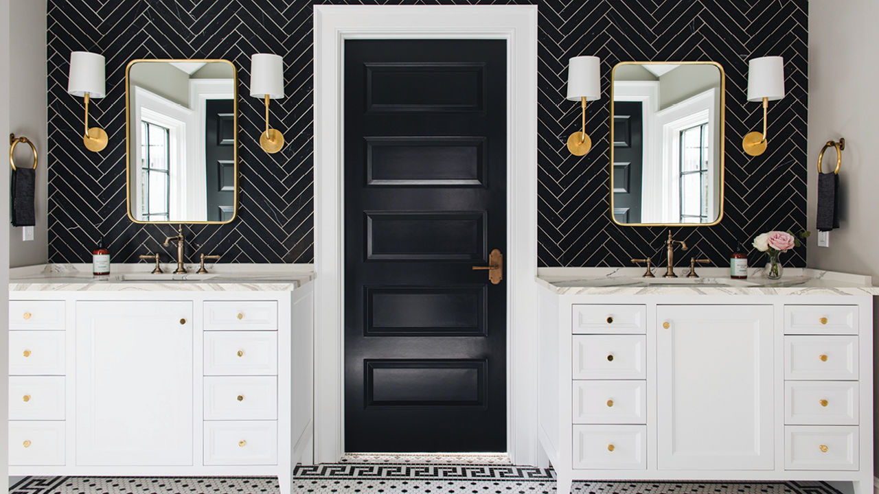 a bold bathroom with two separate vanities with white cabinets and white quartz countertops with a black herringbone-tile backsplash, gold accents in the mirror, hardware, and lighting, with a black door in the center of the vanities. 