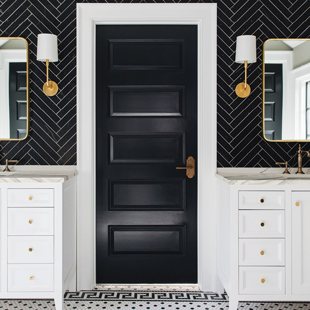 a bold bathroom with two separate vanities with white cabinets and white quartz countertops with a black herringbone-tile backsplash, gold accents in the mirror, hardware, and lighting, with a black door in the center of the vanities. 