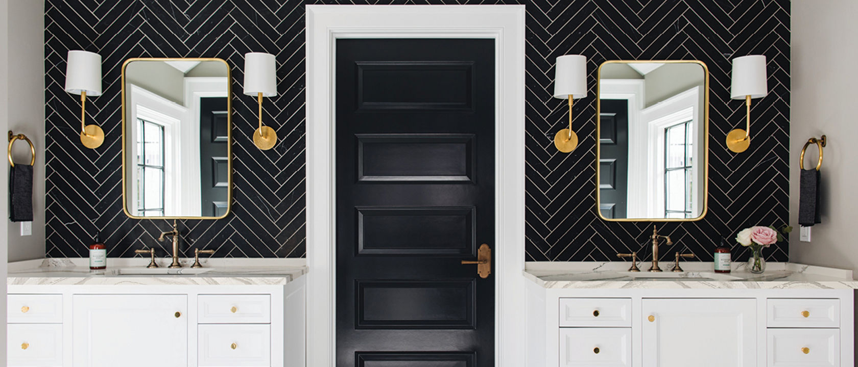 a bold bathroom with two separate vanities with white cabinets and white quartz countertops with a black herringbone-tile backsplash, gold accents in the mirror, hardware, and lighting, with a black door in the center of the vanities. 