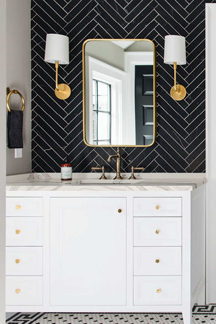 a bold bathroom with two separate vanities with white cabinets and white quartz countertops with a black herringbone-tile backsplash, gold accents in the mirror, hardware, and lighting, with a black door in the center of the vanities. 