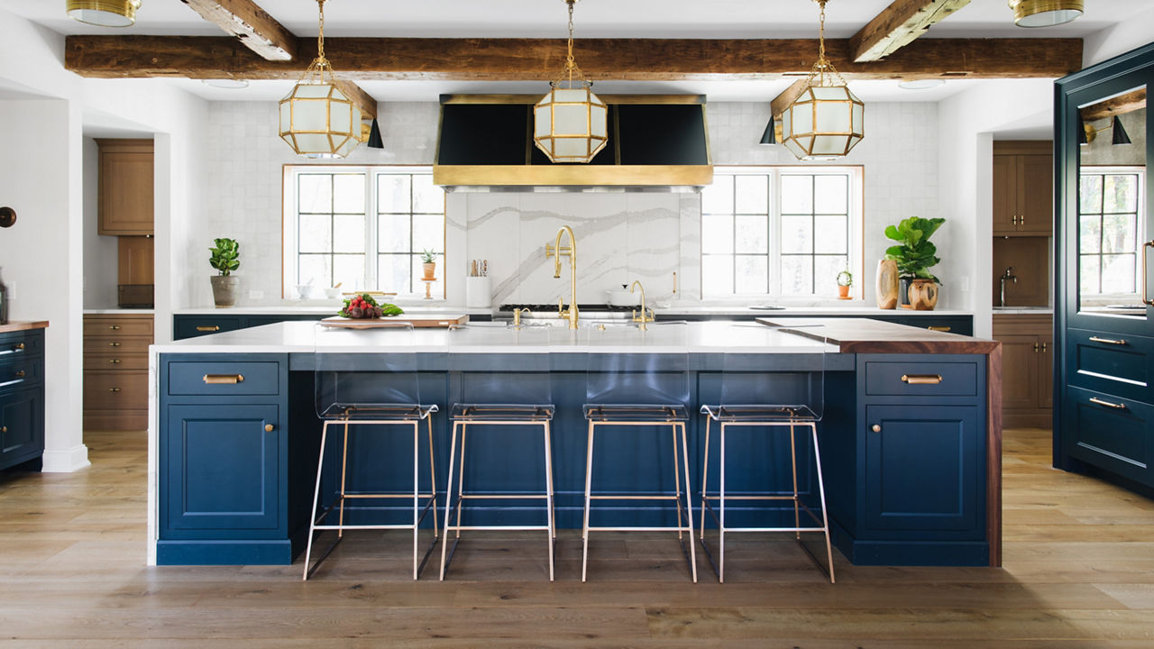 A grand kitchen with a blue center island topped with white quartz countertops, matching quartz backsplash, wooden beams on the ceiling, black and gold hood over the range, and lots of natural light