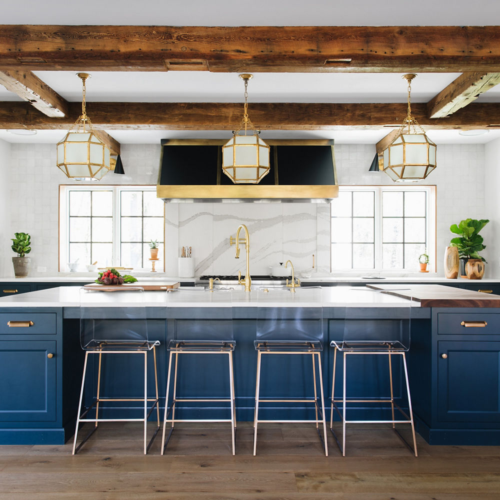 A grand kitchen with a blue center island topped with white quartz countertops, matching quartz backsplash, wooden beams on the ceiling, black and gold hood over the range, and lots of natural light