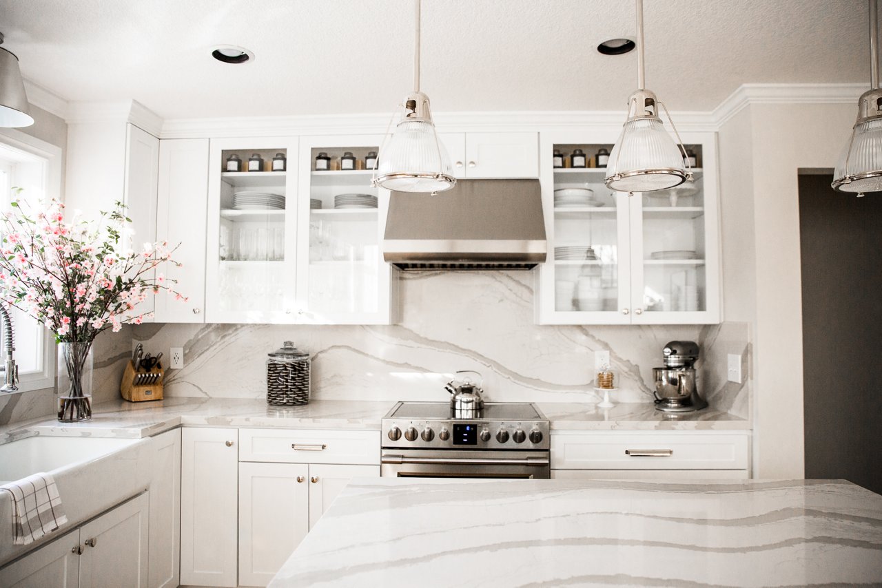 A clean white kitchen with white cabinets, white quartz countertops, stainless steel appliances, lots of natural lighting and overhead pendant lights.