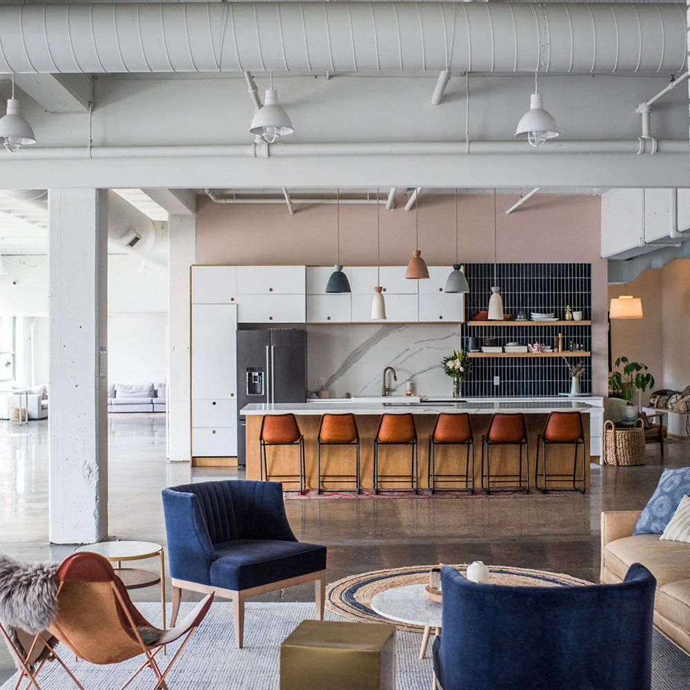 An industrial style workspace featuring a sitting area with cobalt blue chairs and a couch with a glamorous kitchen in the background.