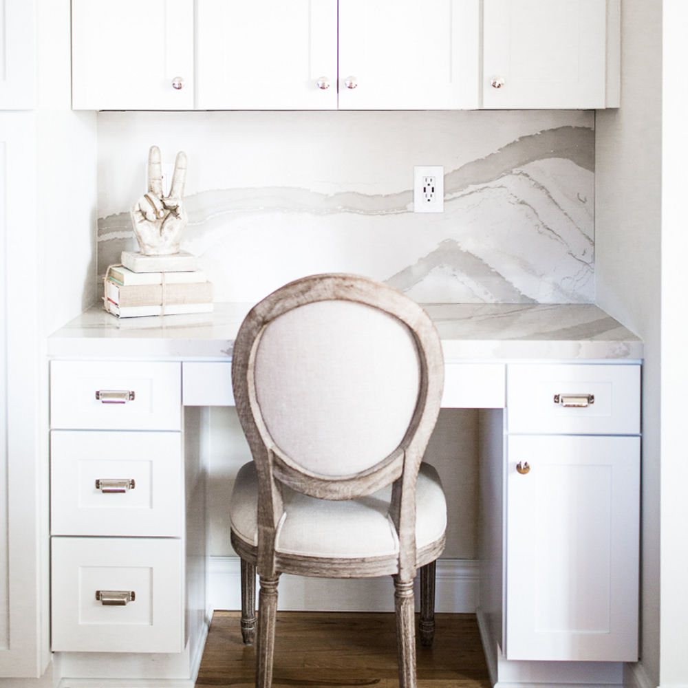 A white kitchen featuring a white chair at a counter with a Cambria Brittanicca Warm quartz countertop and backsplash.