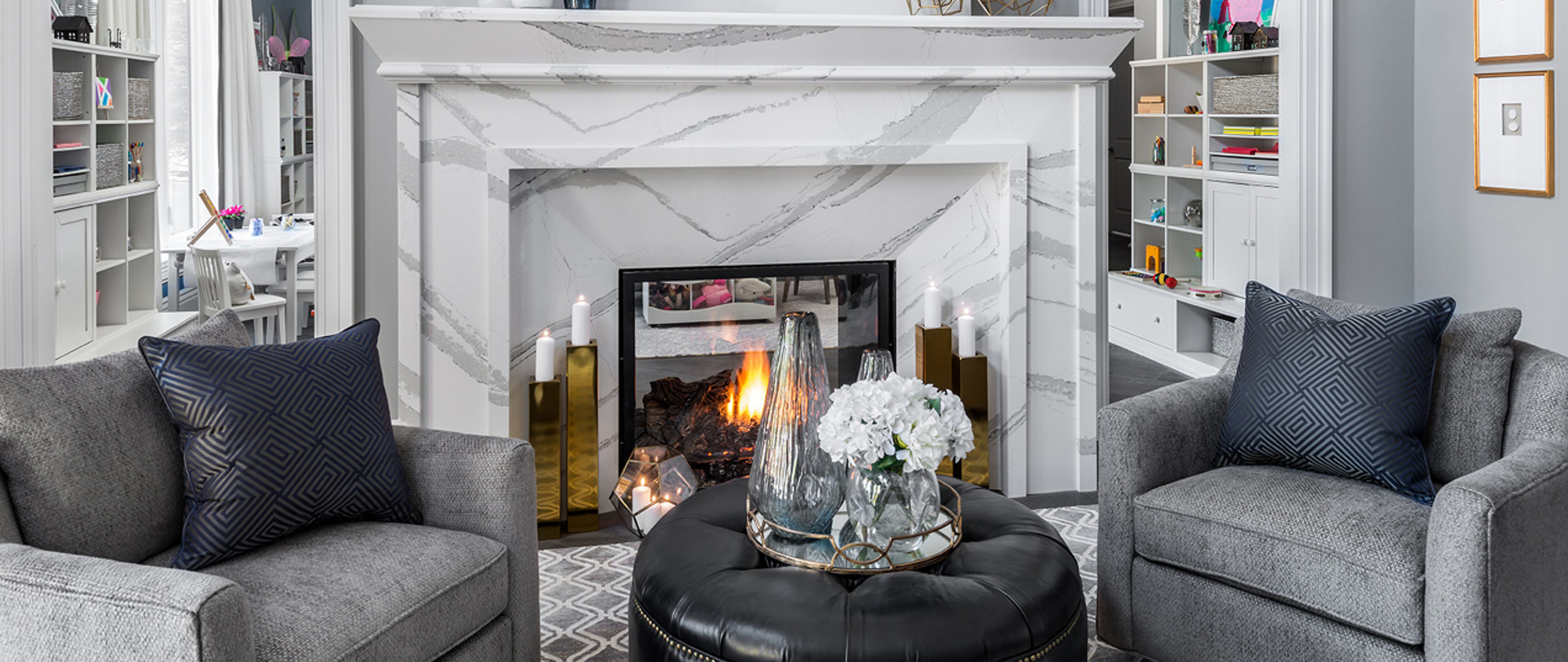 a stunning living room with cool tones, a white and gray veined quartz fireplace, gray armchairs, a black ottoman with a serving tray on top, and gold, blue and white accents throughout. 