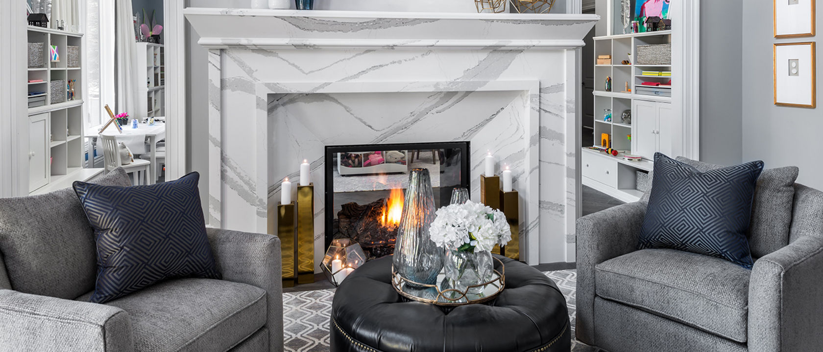 a stunning living room with cool tones, a white and gray veined quartz fireplace, gray armchairs, a black ottoman with a serving tray on top, and gold, blue and white accents throughout. 
