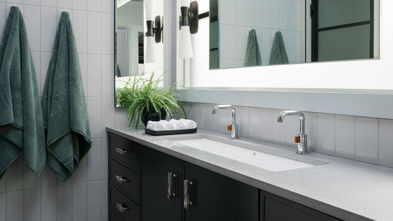 A modern bathroom with a black vanity topped with gray quartz countertops, a double sink, backlit mirror, and two green robes hanging on the gray tiled walls.