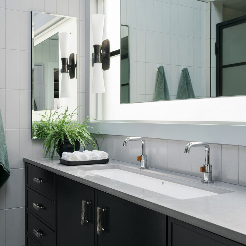 A modern bathroom with a black vanity topped with gray quartz countertops, a double sink, backlit mirror, and two green robes hanging on the gray tiled walls.