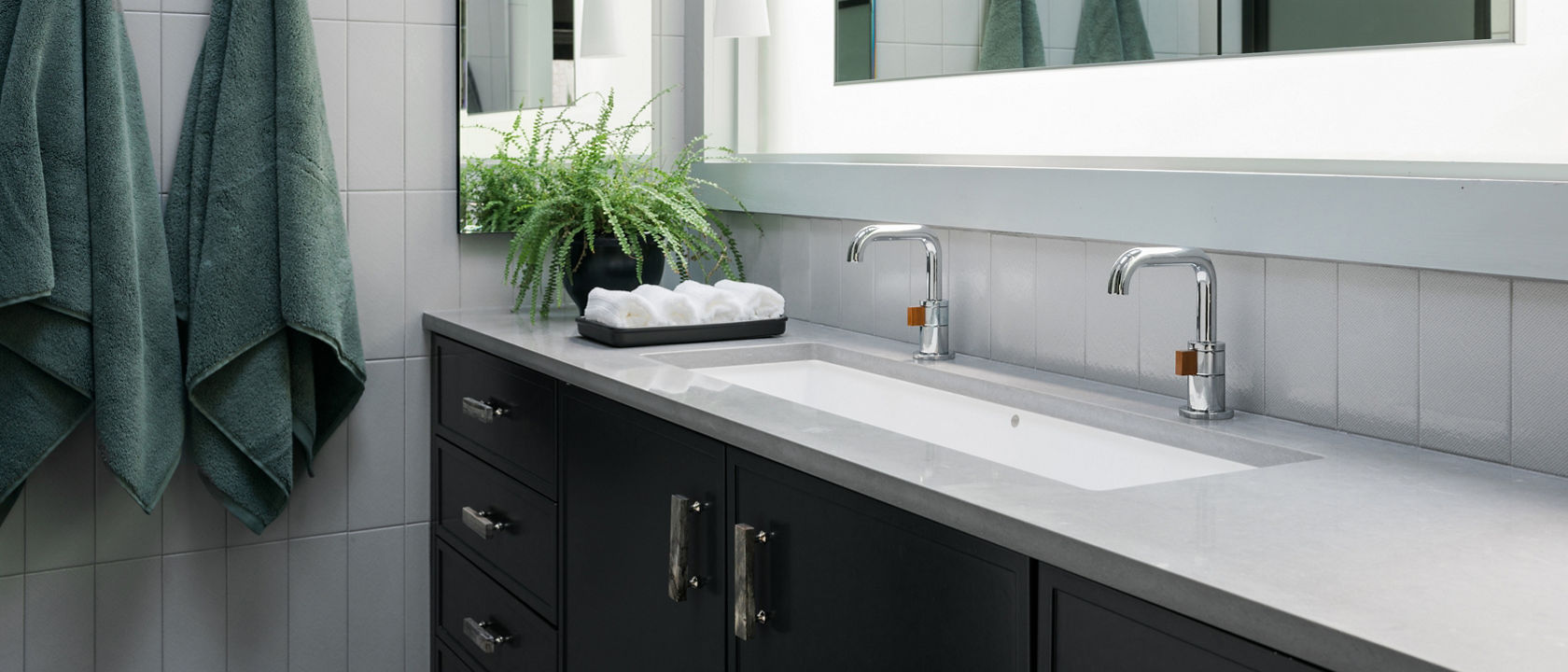 A modern bathroom with a black vanity topped with gray quartz countertops, a double sink, backlit mirror, and two green robes hanging on the gray tiled walls.