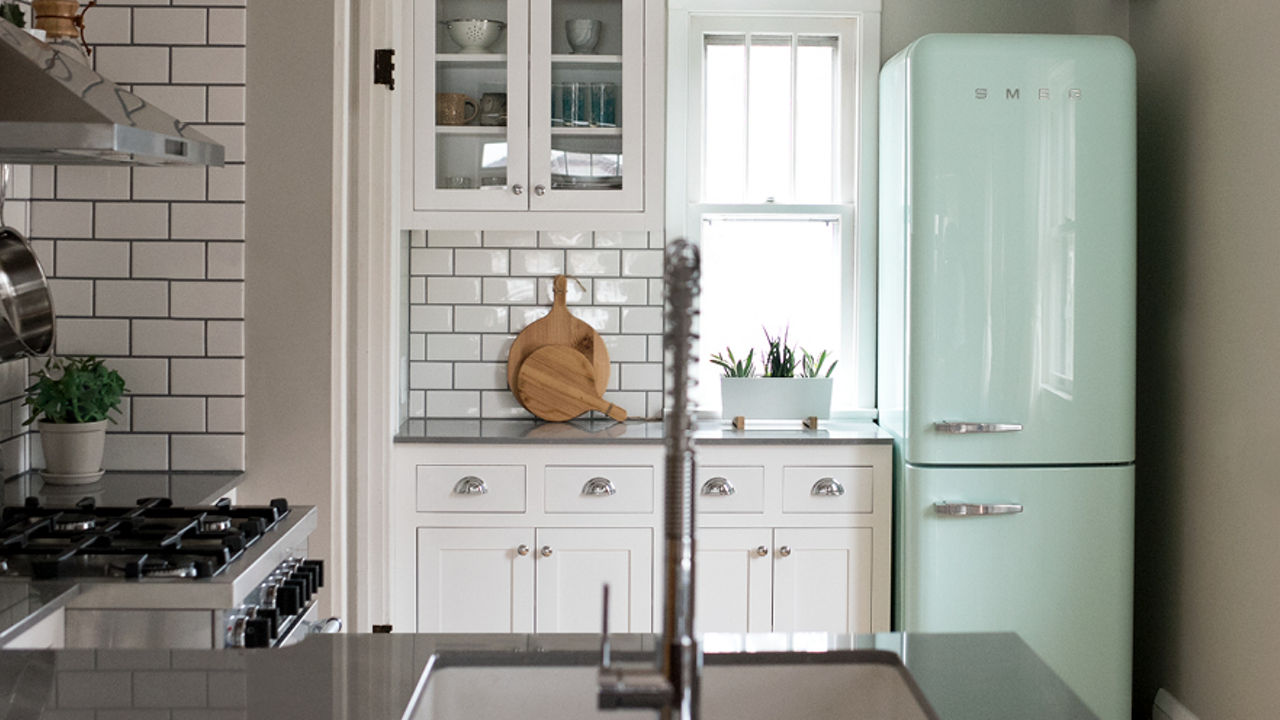 A galley kitchen with white subway tiles, white upper and lower cabinets, a mint green Smeg fridge, gray quartz  countertops, and a small window by the fridge. 