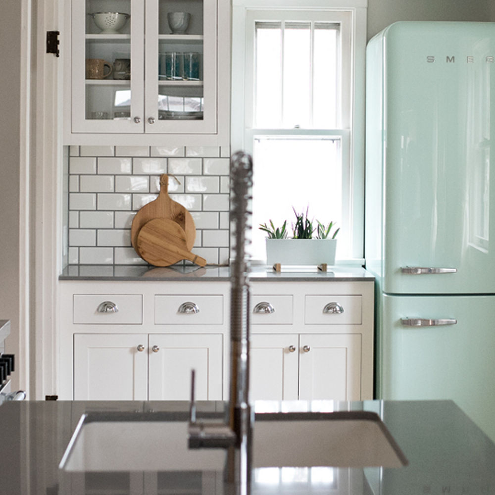 A galley kitchen with white subway tiles, white upper and lower cabinets, a mint green Smeg fridge, gray quartz  countertops, and a small window by the fridge. 