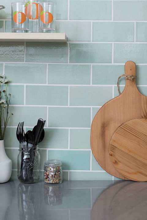 a close up view of a green tile backsplash with gray quartz countertops, open shelving, a rounded cutting board, some books and other miscellaneous decorations