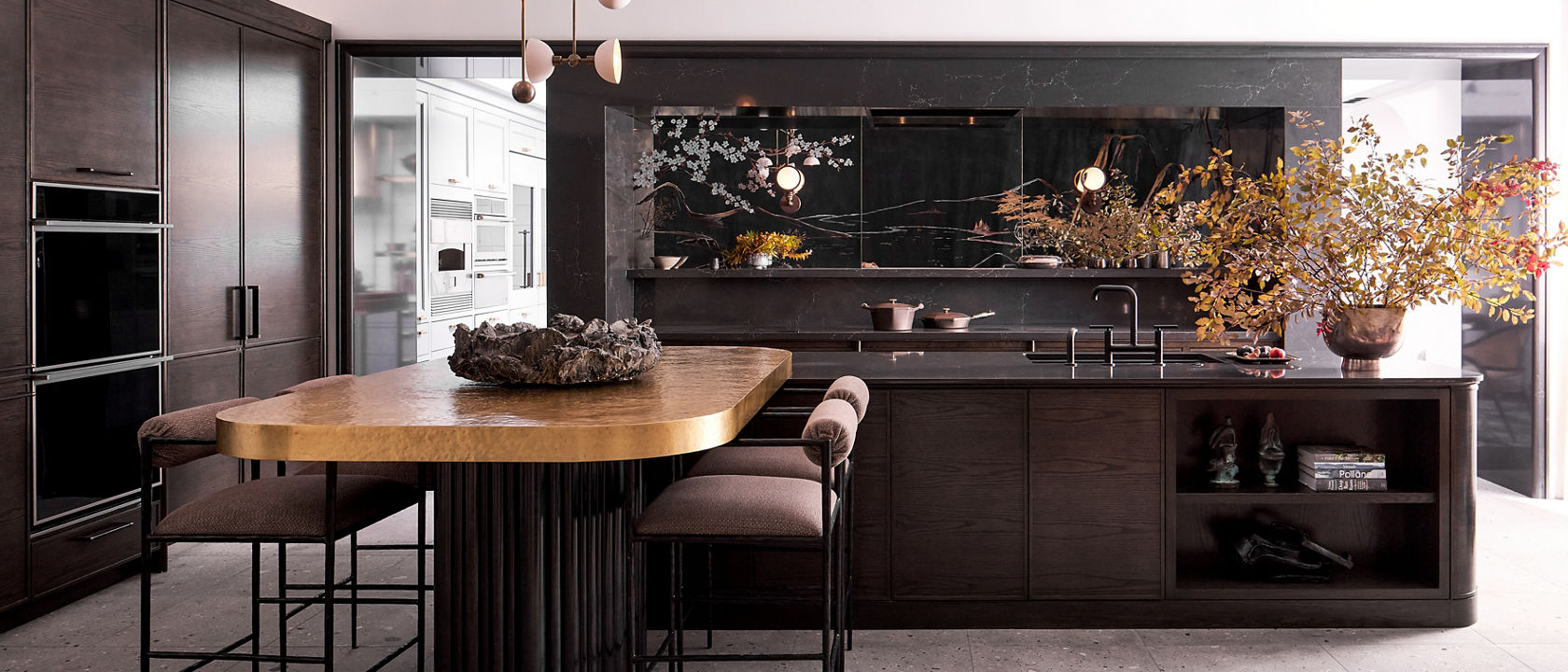 A dark and sleek modern kitchen with dark oak cabinets, tile flooring, Charlestown quartz countertops, column refrigerator, stacked ovens and a wooden dining table with plush seating and gorgeous modern light fixture above. 