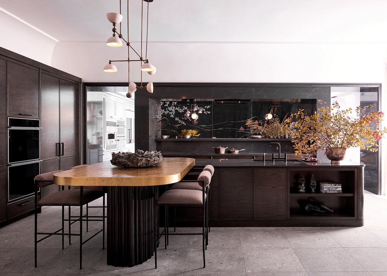 A dark and sleek modern kitchen with dark oak cabinets, tile flooring, Charlestown quartz countertops, column refrigerator, stacked ovens and a wooden dining table with plush seating and gorgeous modern light fixture above. 