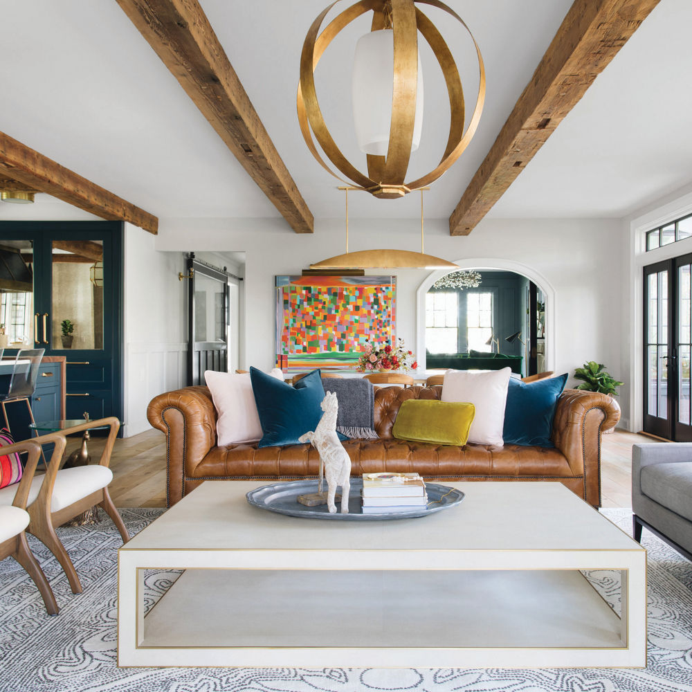 a living room with a leather sofa, white coffee table, wooden beams on the ceiling, a gold chandelier, an ottoman, and two chairs.