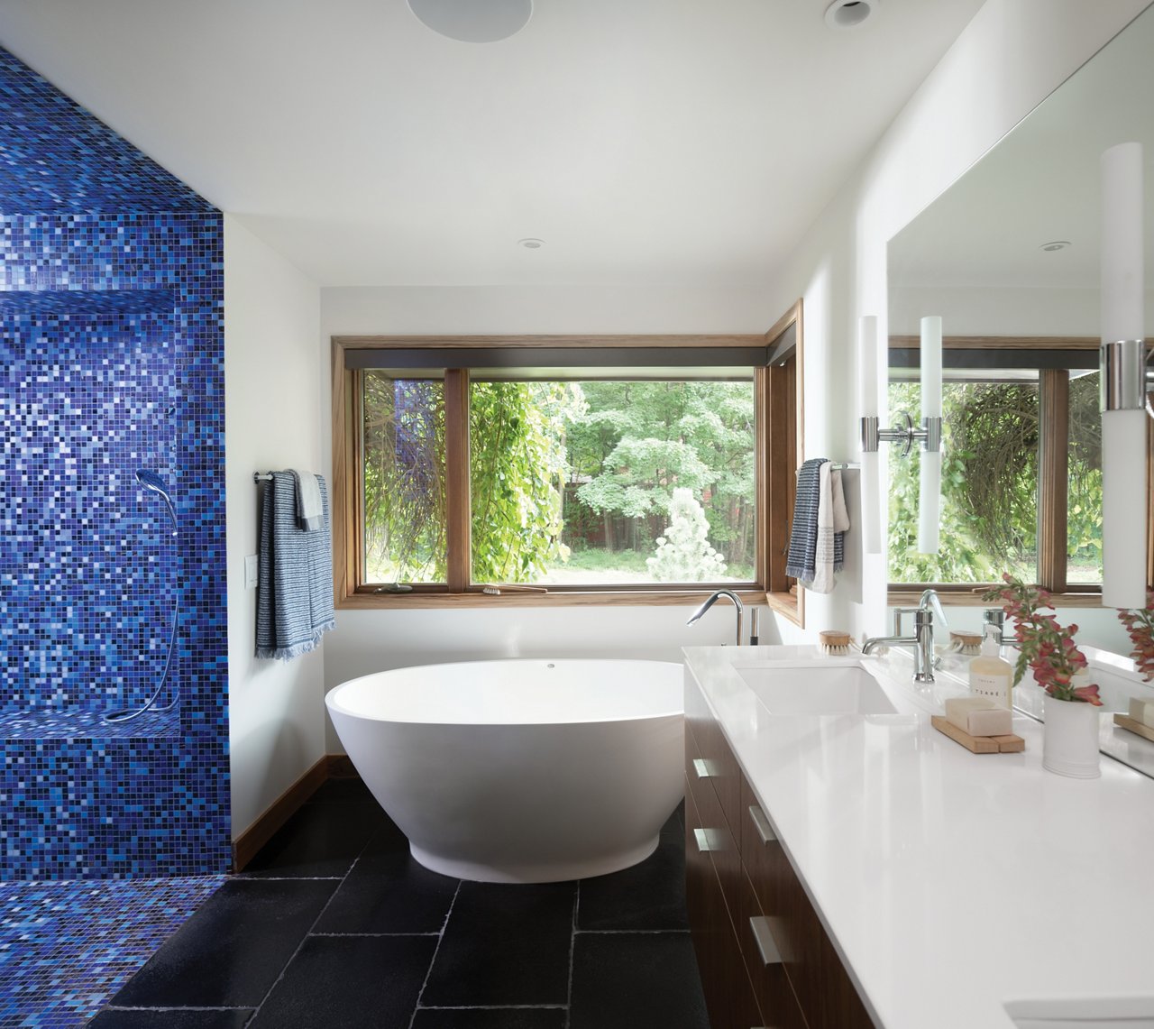 a mid-century modern bathroom with black tile flooring, a blue mosaic tiled shower, a large soaking tub, and a vanity topped with white quartz countertops.