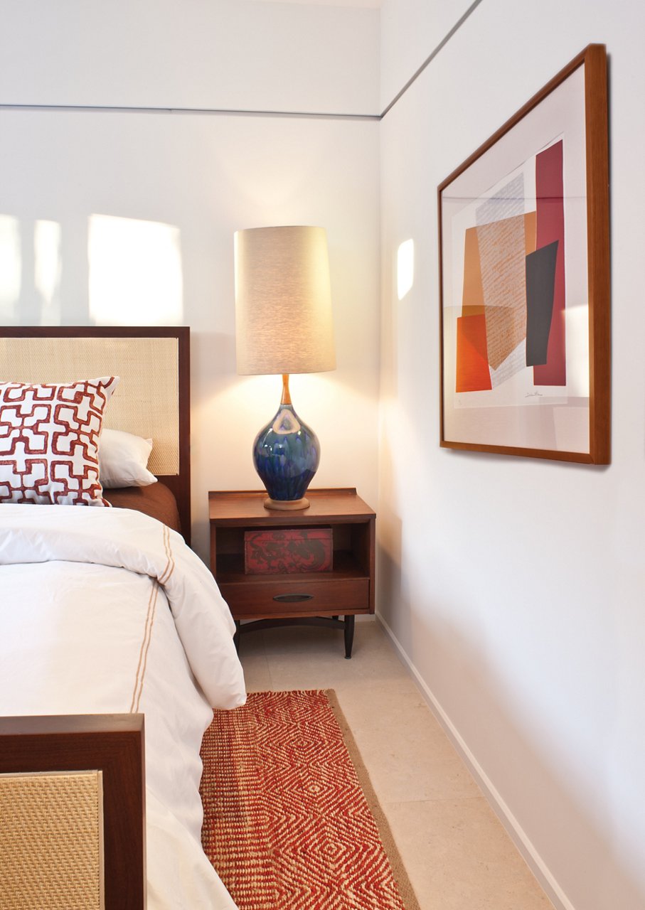 a mid-century modern bedroom with white walls, a wooden framed bed with white bedding, a fun lamp, and art on the wall.