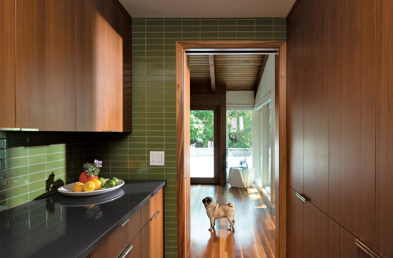 A mid-century modern pantry with oak cabinets, black quartz countertops, green tile backsplash, and a frame doorway.
