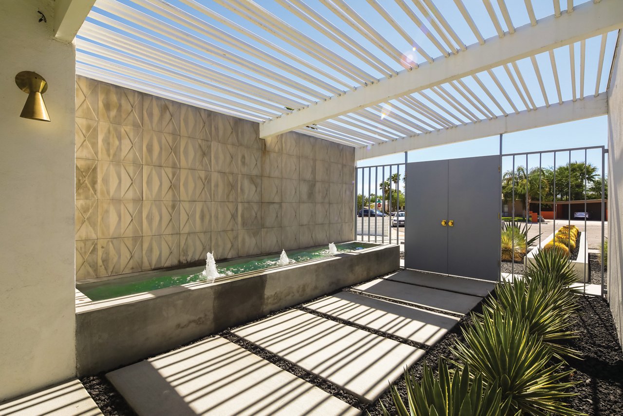 A mid-century modern front car park with a white pergola to provide shade, desert landscaping, and a fountain. 