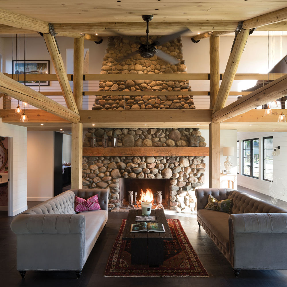 A cabin-style great room with a stone fireplace, wooden beams, and 2 gray couches.